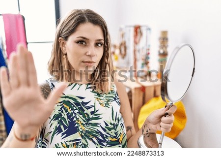 Young hispanic customer woman looking earrings on the mirror at store with open hand doing stop sign with serious and confident expression, defense gesture 