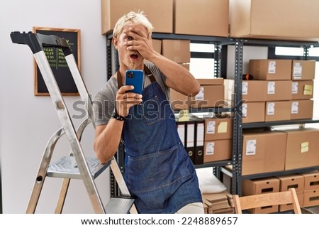 Young blond man using smartphone working at storehouse peeking in shock covering face and eyes with hand, looking through fingers with embarrassed expression. 