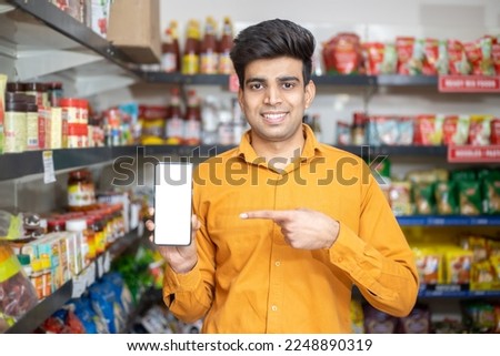 Portrait of happy handsome young Indian man showing smart phone with blank display screen to put advertisement at grocery shop or supermarket, Closeup. Royalty-Free Stock Photo #2248890319