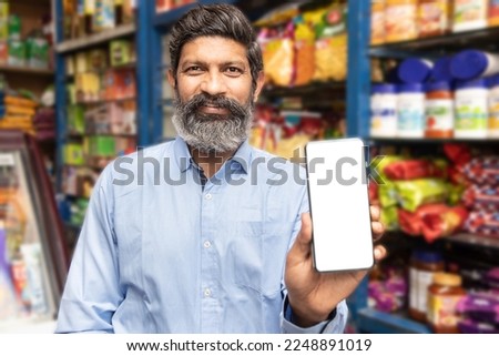 Portrait of happy mature beard Indian man showing smart phone with blank display screen to put advertisement at grocery shop or supermarket, Closeup. Royalty-Free Stock Photo #2248891019