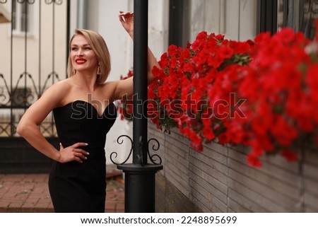 Outdoor street style portrait of blonde 35+ woman with red lips in black  sleeveless dress posing next to blooming red flowers hanging on wall.