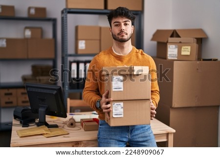 Hispanic man with beard working at small business ecommerce holding packages relaxed with serious expression on face. simple and natural looking at the camera. 