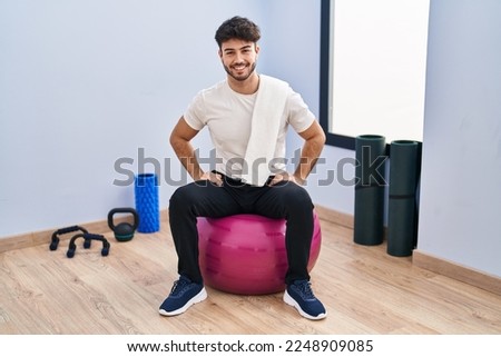 Hispanic man with beard sitting on pilate balls at yoga room with a happy and cool smile on face. lucky person. 