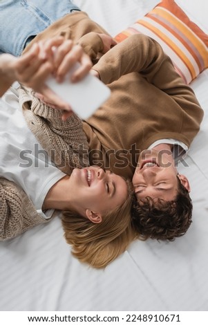 top view of happy couple of travelers taking selfie on blurred smartphone on bed in hotel