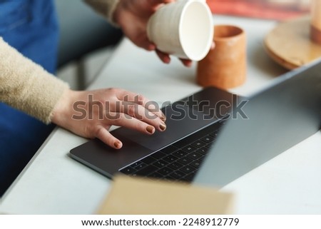 Potter entrepreneur using laptop in workshop, hands only, holding ceramic cup in hands, takes orders online.