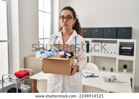 Young hispanic woman holding box with medical items puffing cheeks with funny face. mouth inflated with air, catching air. 