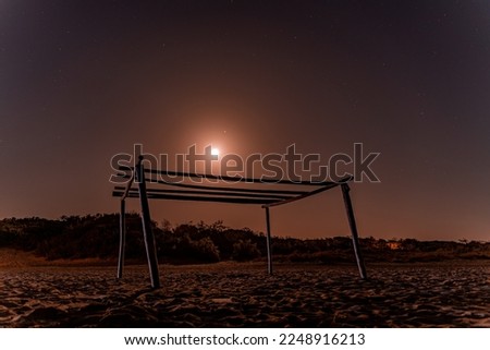 Beautiful photo of a wooden frame built on the beach. Above the wooden poles you can see a beautiful starry sky and a full moon. Tuscany, Italy.