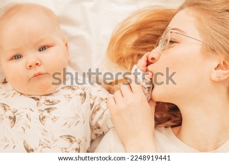 Cheerful young mother kiss fingers on palm of little baby lying on bed closeup. Home family photo of woman and cute infant child resting together. Concept of maternal affection and childcare.