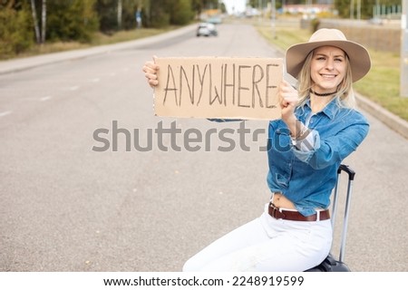 Funny, laughing, cheerful, carefree blond woman in cowgirl hat and jeans shirt hitchhiking by road with cardboard plate anywhere sit on baggage suitcase and wait for car share traffic. Tourist goal