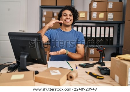 Hispanic man with curly hair working at small business ecommerce gesturing with hands showing big and large size sign, measure symbol. smiling looking at the camera. measuring concept. 