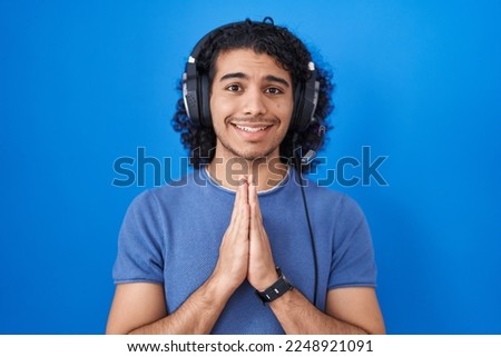 Hispanic man with curly hair listening to music using headphones praying with hands together asking for forgiveness smiling confident. 