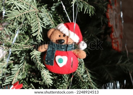 Horizontal Christmas photo of a Christmas tree decoration in the form of a soft bear cub in a scarf,  Santa's hat with a heart, and a button on the body. In the background is a Christmas tree branch. 