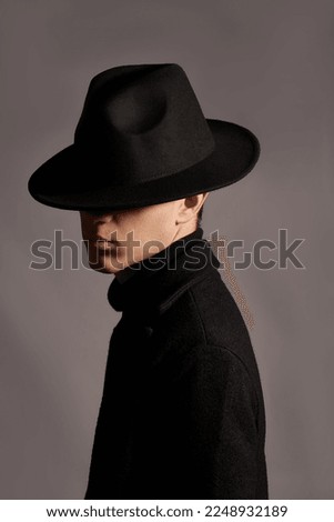 Cropped close-up shot of a man in a black wide-brimmed fedora. A young man in black clothes and a felt hat covering face is posing on a dark background. Side view.               Royalty-Free Stock Photo #2248932189