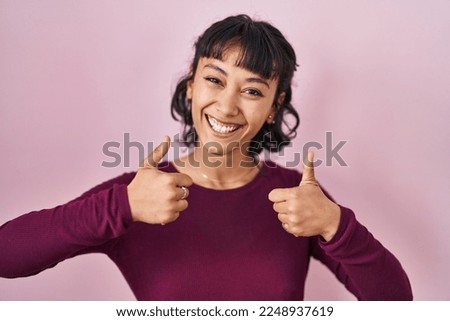 Young beautiful woman standing over pink background success sign doing positive gesture with hand, thumbs up smiling and happy. cheerful expression and winner gesture. 