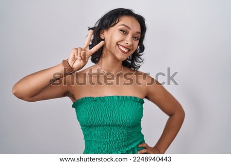 Young hispanic woman standing over isolated background smiling looking to the camera showing fingers doing victory sign. number two. 