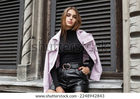 Stylish beautiful young woman model in fashionable black clothes with a pink jacket sits near a vintage building. Trendy elegant pretty girl 