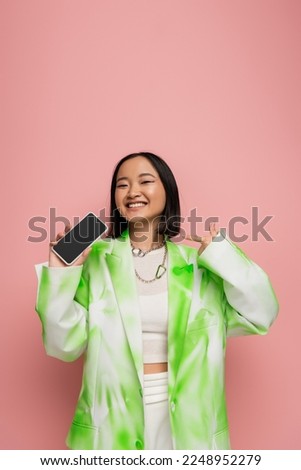 young asian woman in trendy jacket and necklaces pointing at smartphone with blank screen isolated on pink
