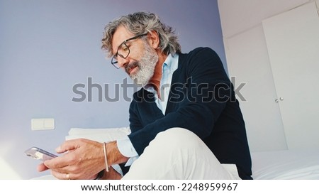 Stylish good looking mature man is sitting on bed and using smartphone
