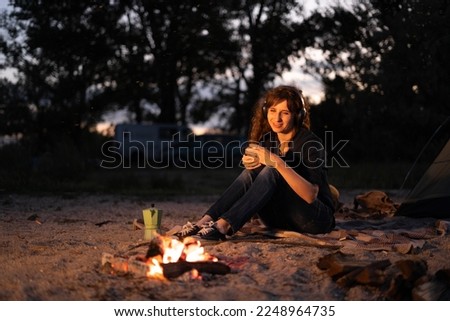 Woman with coffee warming near campfire in forest listening music in headphones. Copy space