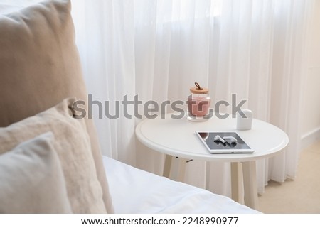 Bedside table with tablet computer, earphones and candle in light bedroom