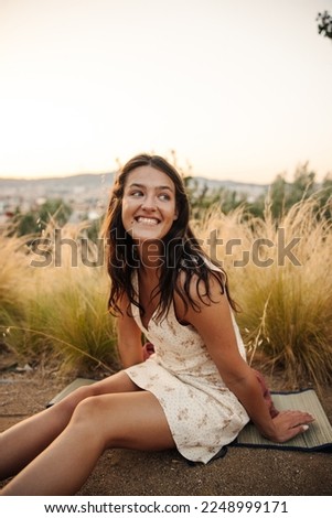 Cheerful young caucasian brunette woman smiles with teeth, looks away sitting outdoors. Girl wears white sundress on summer day. Relaxation concept Royalty-Free Stock Photo #2248999171