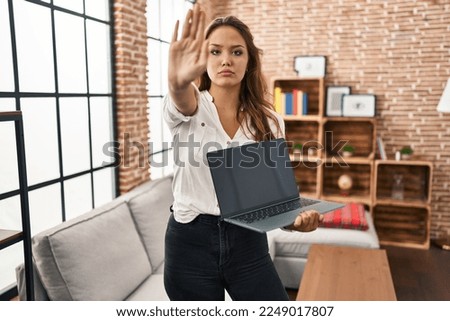 Young hispanic woman using laptop showing blank screen at home with open hand doing stop sign with serious and confident expression, defense gesture 
