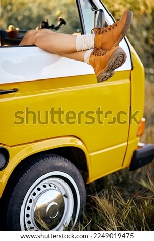 Cropped female in orange boots travelling on yellow mini van, in field, legs peeped out from retro van, close-up photo of legs, exploring new places in countryside, trip. vacation, people lifestyle