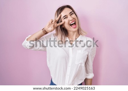 Young beautiful woman standing over pink background doing peace symbol with fingers over face, smiling cheerful showing victory 