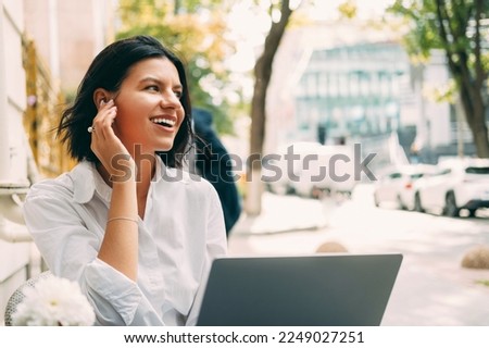 Cheerful woman is working at the laptop outdoors at a cafe table on the street and wearing wireless ear pods. Royalty-Free Stock Photo #2249027251
