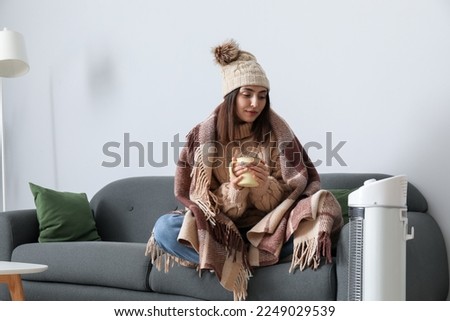 Frozen young woman with cup of tea warming near electric heater at home