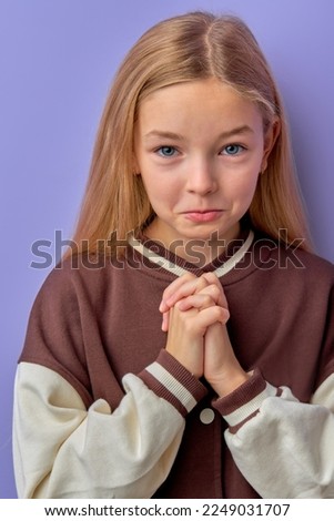 Forgive me. Little blonde teen kid child girl praying, looking at camera and making wish, asking with hopeful imploring expression, begging apology isolated on purple background. Children emotions