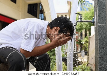 Muslim kid wearing koko shirt and sarong is sitting and cleansing face by hands using water outside the mosque, procedure of ablution before praying salah.  Royalty-Free Stock Photo #2249032353