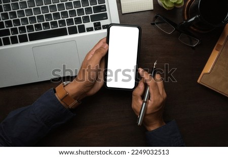 Above view of man hands holding smart phone with white empty screen on wooden desk.