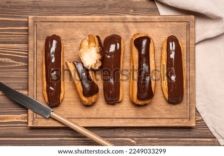 Cutting board with tasty chocolate eclairs on wooden background Royalty-Free Stock Photo #2249033299