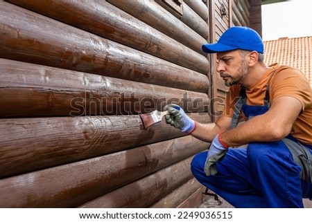 Man protecting mountain house wooden walls by applying protective varnish or paint with brush Royalty-Free Stock Photo #2249036515