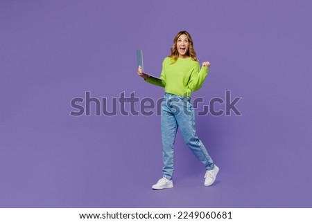 Full body young fun side view IT woman 30s she wears casual green knitted sweater hold use work on laptop pc computer walk do winner gesture isolated on plain pastel purple background studio portrait