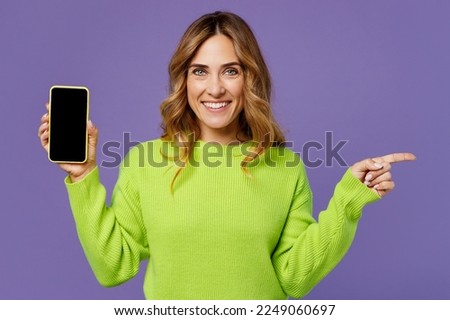 Young woman she wearing green knitted sweater hold in hand use mobile cell phone with blank screen workspace point index finger aside on area isolated on plain pastel purple background studio portrait