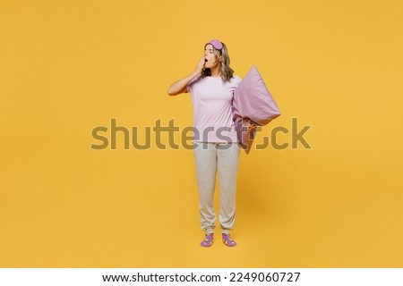 Full body tired young woman she wears purple pyjamas jam sleep eye mask rest relax at home cover mouth with hand yawn hold pillow isolated on plain yellow background studio portrait. Night nap concept Royalty-Free Stock Photo #2249060727