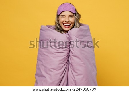 Fun young happy cheerful woman she wearing pyjamas jam sleep eye mask wrapped in duvet blanket wink rest relax at home isolated on plain yellow background studio portrait. Good mood night nap concept