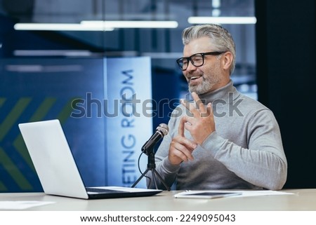 Senior gray-haired man sitting at a desk in an office with a microphone and laptop. Talks on a video call. Conducts interviews, business meetings, webinars, online meetings. Shows, counts on fingers.