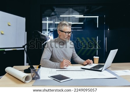 Senior gray-haired man architect, engineer, designer sits in the office at the table, works on a laptop, drawings. Makes notes.