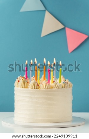 Birthday cake with white cream cheese frosting decorated with multicolored lit candles on a blue background. Happy Birthday concept. 
Tradition of making a wish while blowing out candles on a cake