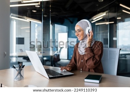 Successful modern businesswoman working inside office with laptop, Muslim woman wearing hijab and headphones listening to audio books and podcasts at workplace, satisfied and successful woman. Royalty-Free Stock Photo #2249110393