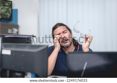 A tired office employee rubbing his tired eyes after a tasking day at his desk job. Eye fatigue from overtime and working long hours looking into the computer screen at the office. Royalty-Free Stock Photo #2249176033