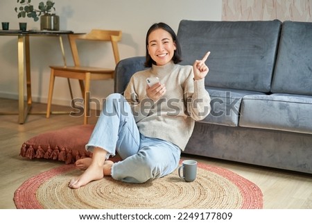 Portrait of modern young korean woman sitting on floor, pointing finger at copy space, showing banner or advertisement, holding smartphone as using app.