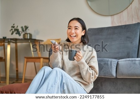 Portrait of beautiful asian girl sitting at her home and watching tv, holding remote, smiling and laughing, feeling comfort and warmth at her apartment. Royalty-Free Stock Photo #2249177855