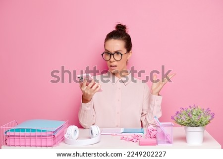 Shocked woman, watch scary content on the smartphone, with unexpected expression, writes important notes on the notebook, isolated on pink wall