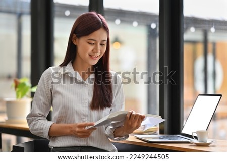Young Asian woman in smart casual reading a book while sitting on the counter bar with Empty of screen laptop.