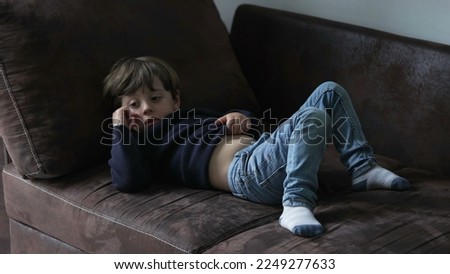 Bored child lying on sofa at home watching TV screen off camera. One little boy feeling boredom