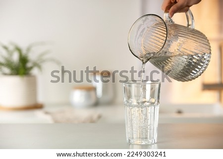 Woman pouring water from jug into glass at white table in kitchen, closeup. Space for text Royalty-Free Stock Photo #2249303241